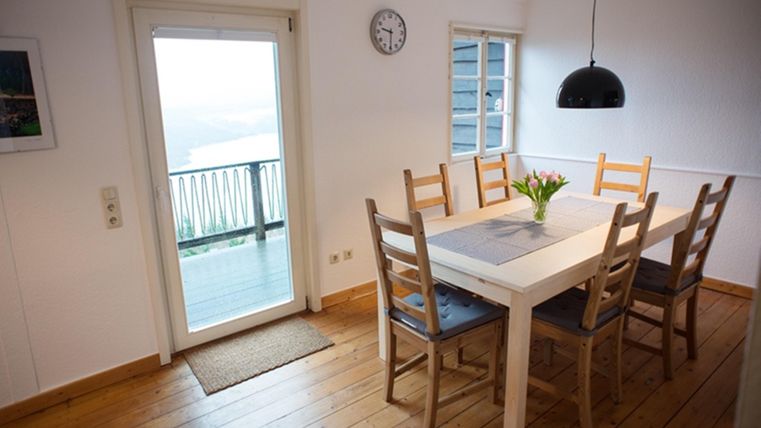 Ein helles Esszimmer mit einem weißen Tisch und Holzstühlen. Eine Blume in der Mitte des Tisches und ein Zugang zu einem Balkon mit Aussicht.