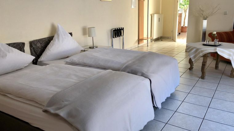 Ein gemütliches Zimmer mit zwei Einzelbetten und hellen, neutralen Farben. Im Hintergrund ist ein Platz mit einem Sofa und viel natürlichem Licht zu sehen.