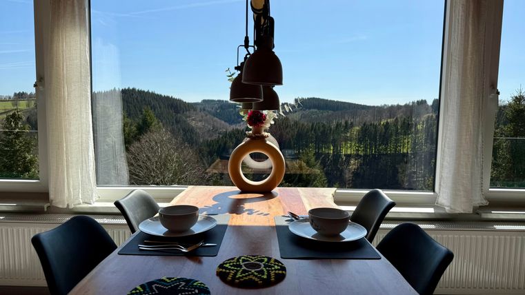 Ein helle Essbereich mit einem Tisch und zwei Schalen. Draußen sieht man eine malerische Landschaft mit Hügeln und einem klaren blauen Himmel.