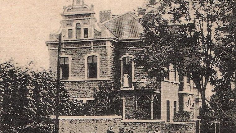 Ein historisches Gebäude mit einem charakteristischen Dach und großen Fenstern. Vor dem Gebäude stehen mehrere Personen in traditioneller Kleidung.