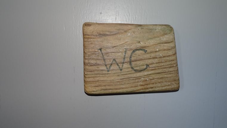 Ein Holzschild mit der Aufschrift "WC". Es ist an einer Wand befestigt.