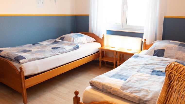 Ein einfaches Zimmer mit zwei Betten, hellen Wänden und einem Fenster. Es gibt eine kleine Nachtischlampe und ein gemütliches Ambiente.