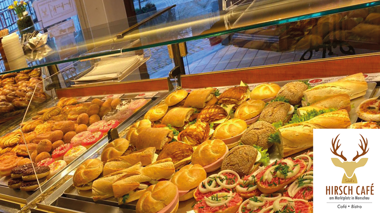 Eine Auswahl an köstlichen Backwaren und Snacks in einer Glasvitrine. Im Hintergrund sind frische Brötchen und verschiedene belegte Brote zu sehen.