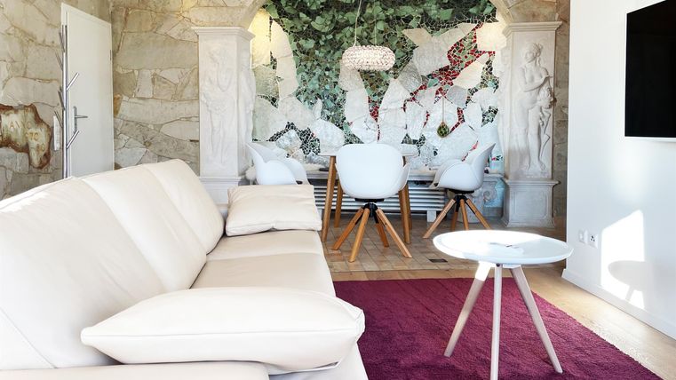 Ein modernes Wohnzimmer mit einem weißen Sofa und einem lila Teppich. Im Hintergrund ist ein schöner Essbereich mit einem kunstvollen Wandbild zu sehen.
