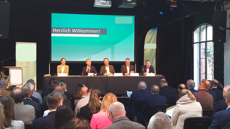 Eine Konferenz mit mehreren Rednern an einem Tisch vor einem Publikum. Im Hintergrund ist ein Bildschirm mit der Aufschrift "Herzlich Willkommen!" zu sehen.