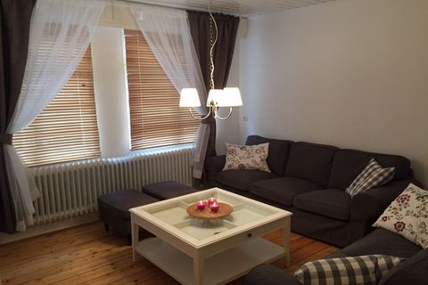 Ein gemütliches Wohnzimmer mit einem grauen Sofa und einem weißen Tisch. Natürliche Lichtverhältnisse durch Fenster mit Jalousien verleihen dem Raum eine helle Atmosphäre.