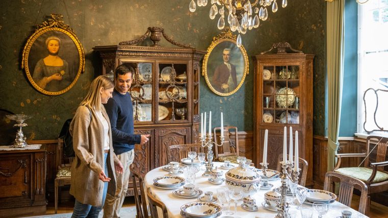 Ein stilvoll eingerichteter Essraum mit einem eleganten Tisch, der mit feinem Geschirr gedeckt ist. Im Hintergrund sind antike Möbel und Portraits an der Wand zu sehen.