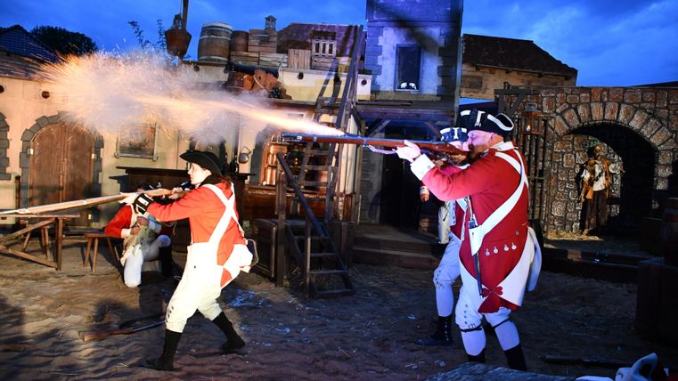 Zwei Soldaten in historischen Uniformen feuern mit Gewehren in einer lebhaften Darbietung. Die Szene spielt in einer alten Kulisse bei Dämmerlicht.