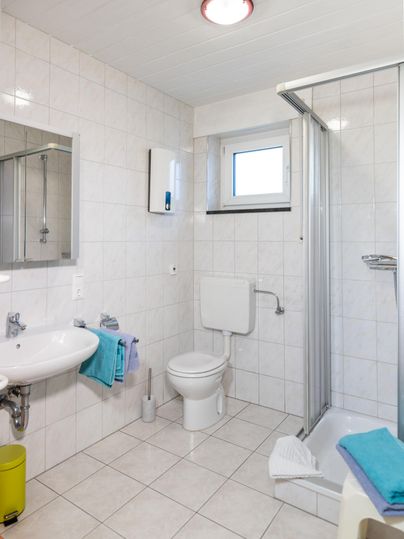Ein helles, modernes Badezimmer mit einer Dusche, Waschbecken und Toilette. Die Wände sind in hellen Fliesen gehalten, und es gibt einige Handtücher in bunten Farben.