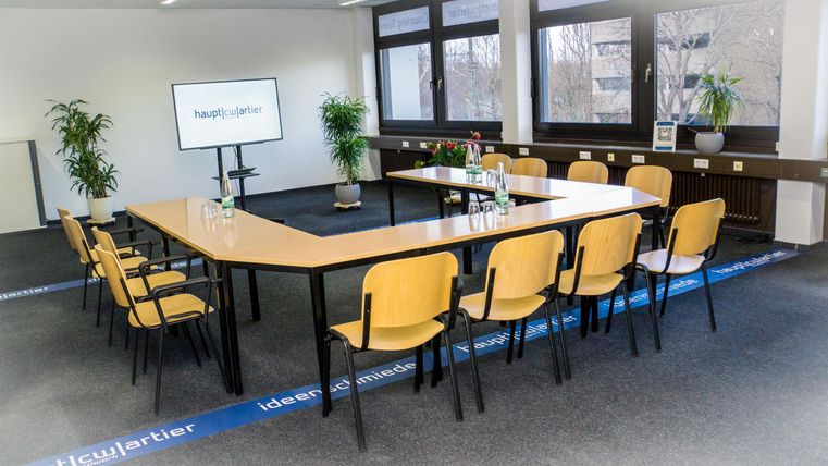 Ein modernes Besprechungszimmer mit einem großen Tisch und gelben Stühlen. Auf einem Bildschirm im Raum ist ein Präsentationstext zu sehen.