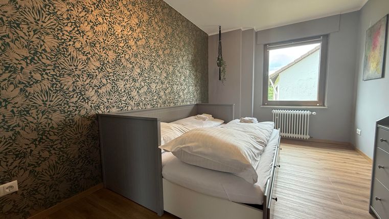 Ein modernes Schlafzimmer mit einem großen Doppelbett und einem ansprechenden Pflanzenmuster an der Wand. Es hat ein Fenster, das Tageslicht hereinlässt, und einen Holzfußboden.