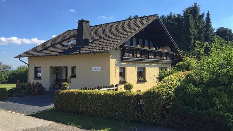 Ein gemütliches Haus mit einem Balkon und einem gepflegten Garten. Im Hintergrund sind Bäume und ein klarer Himmel zu sehen.