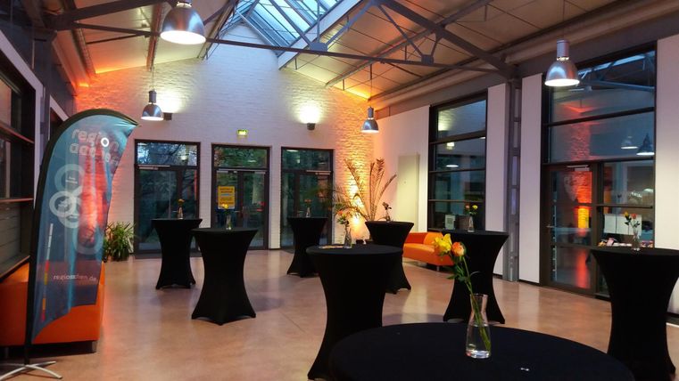 Ein moderner Veranstaltungsraum mit großen Fenstern und stilvollen Stehtischen. Die Beleuchtung schafft eine einladende Atmosphäre für Events.