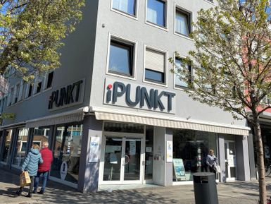 Ein modernes Geschäft mit dem Namen "iPUNKT" in einer städtischen Umgebung. Menschen gehen vorbei und es gibt Bäume, die Schatten spenden.