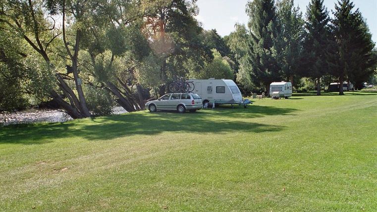 Ein ruhiger Campingplatz am Wasser mit grünen Wiesen. Zwei Wohnmobile stehen unter schattigen Bäumen.