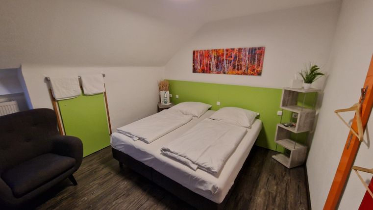 Ein gemütliches Schlafzimmer mit einem Doppelbett und einer grünen Wand. Eine Sitzecke mit einem Sessel und einem modernen Regal sind ebenfalls vorhanden.