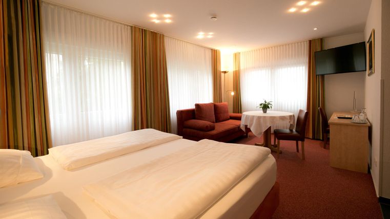 Ein modernes Hotelzimmer mit zwei Betten, einem Sofa und einem Tisch. Das Zimmer hat große Fenster mit Vorhängen und einen Fernseher an der Wand.