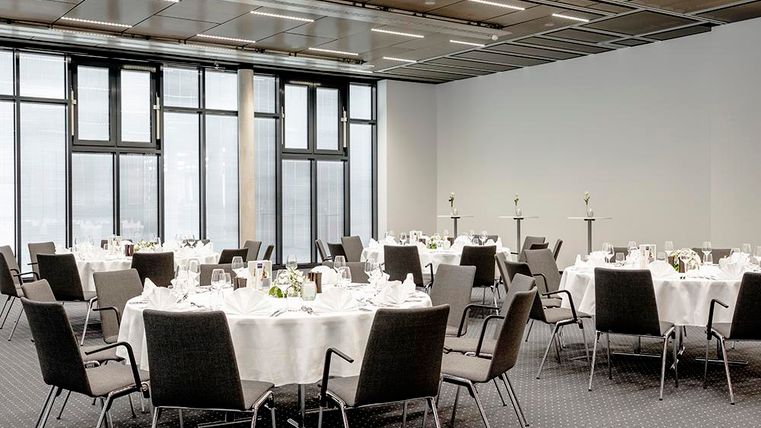 Ein eleganter Raum mit mehreren runden Tischen, die für ein Event eingedeckt sind. Große Fenster lassen viel Licht herein und bieten eine moderne Atmosphäre.