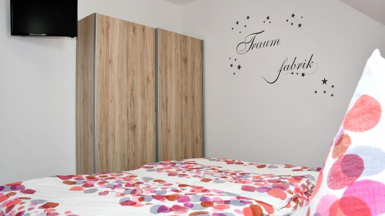 Ein gemütliches Schlafzimmer mit einem Bett und bunten Kissen. An der Wand steht der Schriftzug "Traumfabrik" und es gibt einen Fernseher.