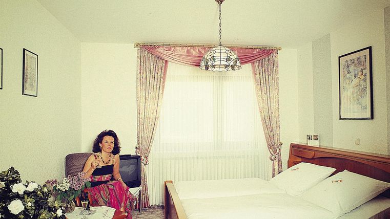 Ein gemütliches Schlafzimmer mit einem Doppelbett und einem Sessel. Eine Frau sitzt entspannt im Raum neben einem Blumenarrangement.