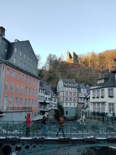 Eine malerische Altstadt mit historischen Fachwerkhäusern und einer Brücke über einen ruhigen Fluss. Im Hintergrund ist eine Burg auf einem Hügel zu sehen.