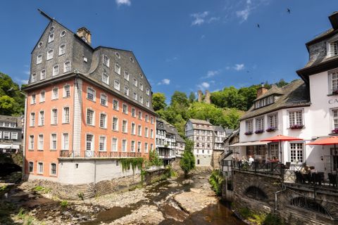 Eine malerische Stadtansicht mit historischen Gebäuden entlang eines kleinen Flusses. Im Hintergrund sind bewaldete Hügel und blauer Himmel zu sehen.