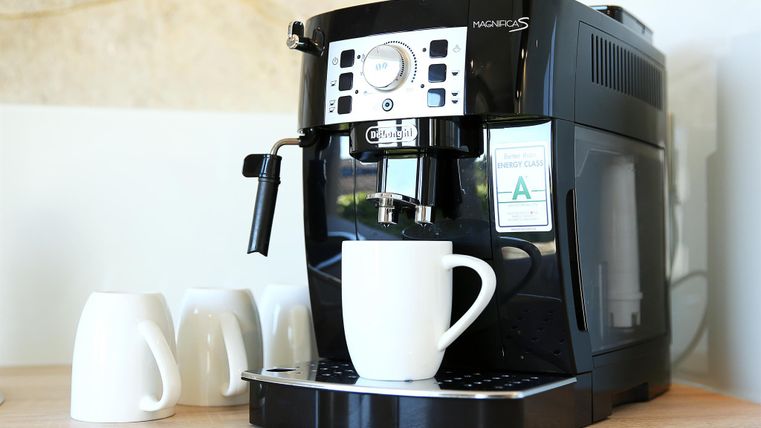 Eine Kaffeemaschine steht auf einem Tisch mit einer weißen Tasse darunter. Daneben stehen mehrere weiße Tassen.