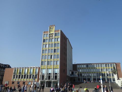 Ein modernes Gebäude mit einer markanten Fassade und einem hohen Turm. Menschen stehen davor und genießen den sonnigen Tag.