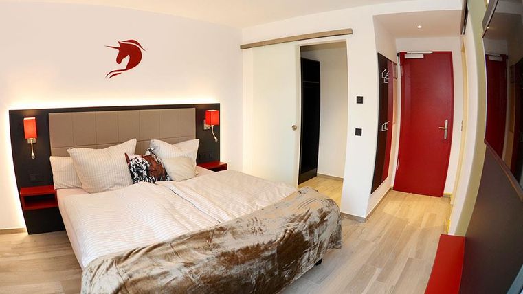 Ein modernes Schlafzimmer mit einem Doppelbett und roten Akzenten. Die Wände sind hell, und es gibt einen großen Spiegel sowie einen Kleiderschrank.
