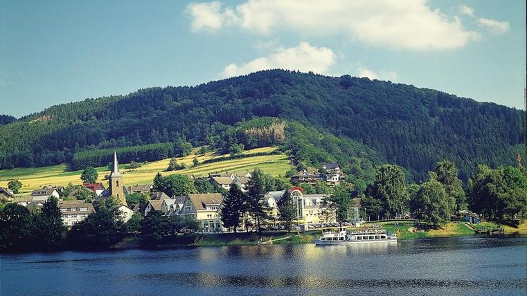 Eine malerische Landschaft mit einem ruhigen See und hügeliger Umgebung. Im Vordergrund sind Häuser und Bäume zu sehen, während im Hintergrund sanfte Berge erheben.
