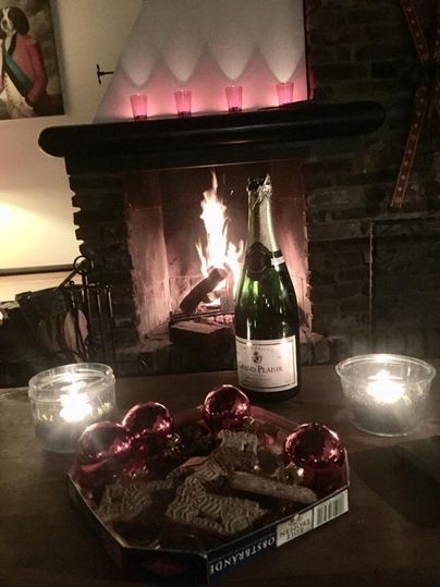 Ein gemütlicher Kamin mit flammendem Feuer und einem Glas Sekt. Auf dem Tisch liegen Lebkuchen und rote Weihnachtskugeln.