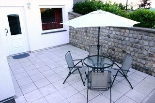 Eine gemütliche Terrasse mit einem runden Glastisch und vier Stühlen. Ein Sonnenschirm sorgt für Schatten und der Bereich ist von einer Steinmauer umgeben.