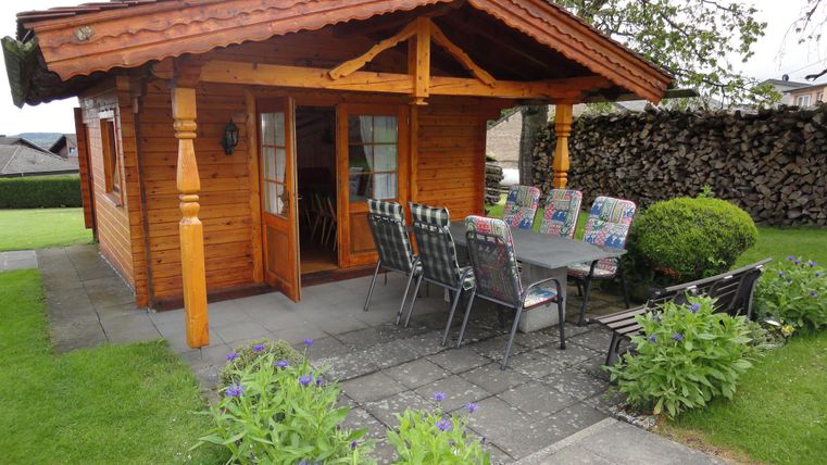 Ein gemütliches Holzhaus mit einer Terrasse und einem Essbereich im Freien. Umgeben von grünem Rasen und bunten Blumen.