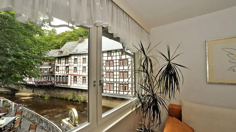 Ein Blick aus dem Fenster auf ein malerisches Gebäude mit Fachwerk und einen ruhigen Fluss. Grüne Bäume und eine gemütliche Sitzecke verleihen der Szene eine einladende Atmosphäre.