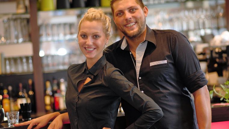 Ein lächelndes Paar steht hinter einer Bar. Der Mann und die Frau tragen schwarze Hemden und wirken freundlich und einladend.