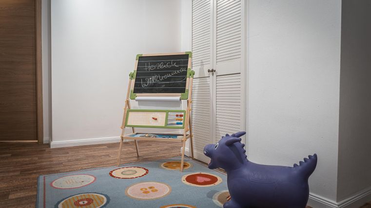 Ein helles Zimmer mit einem Spielteppich und einem kleinen, violetten Hüpfspielzeug. An der Wand steht eine kleine Tafel zum Zeichnen und Schreiben.