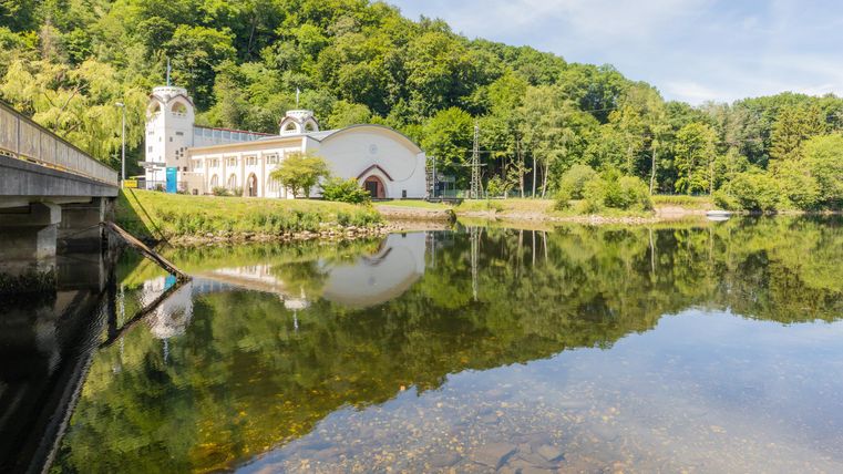 Ein ruhiger See mit klaren Wasserreflexionen und umgebenden grünen Bäumen. Im Hintergrund ist ein historisches Gebäude sichtbar.