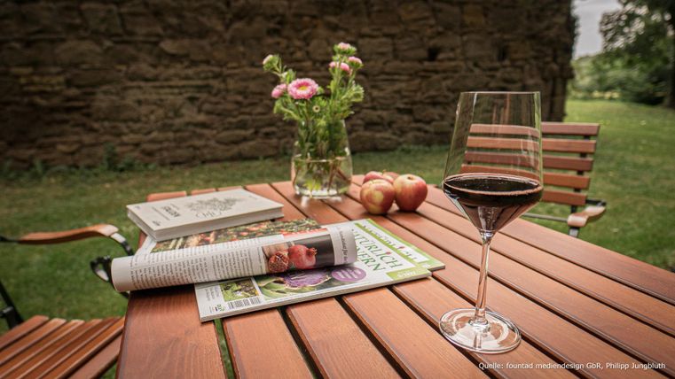Ein hübscher Tisch aus Holz im Freien, dekoriert mit einem Glas Rotwein, Zeitschriften und einer Vase mit Blumen. Im Hintergrund sind grüne Wiesen zu sehen.