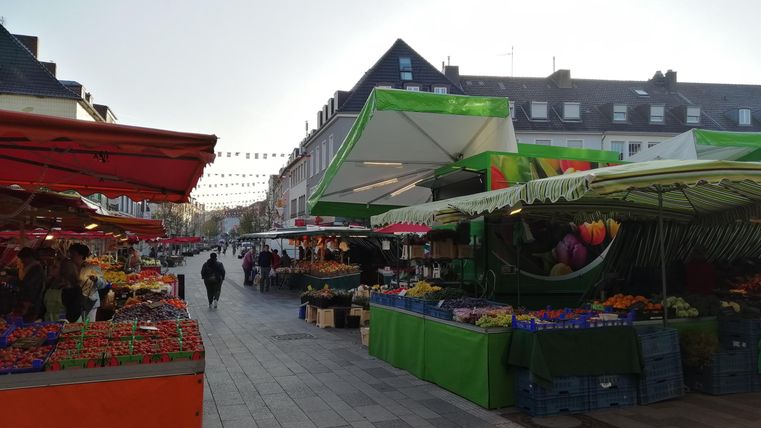 Ein belebter Markt mit bunten Ständen und frischen Produkten. Viele Menschen schlendern entlang der Straßen unter einem klaren Himmel.