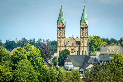 Eine beeindruckende Kirche mit zwei spitzen Türmen steht zwischen Bäumen und Häusern. Der Himmel ist klar und blau, was die Szene lebendig wirkt.