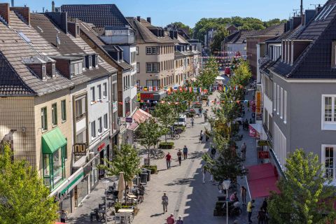Eine belebte Einkaufsstraße mit Bäumen und vielen Menschen. Farbenfrohe Fahnen schmücken die Straße und laden zum Bummeln ein.