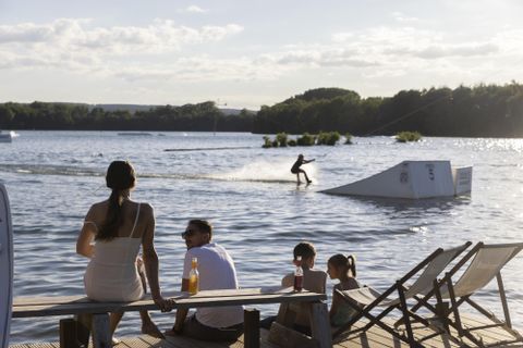 Eine Gruppe von Menschen sitzt am Ufer eines Gewässers. Im Hintergrund fährt jemand auf einem Wassersportgerät über die Wasseroberfläche.