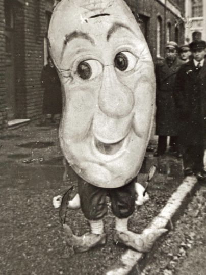 Eine Person in einem großen Maskenkostüm lächelt. Im Hintergrund stehen mehrere Menschen in historischer Kleidung.