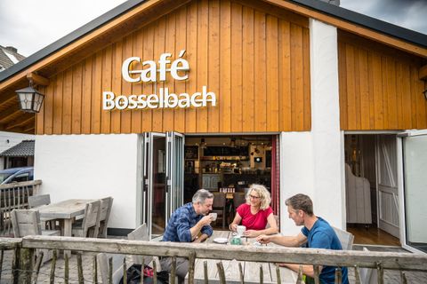 Ein gemütliches Café mit dem Namen "Café Bosselbach". Drei Personen genießen ihre Zeit draußen an einem Tisch.
