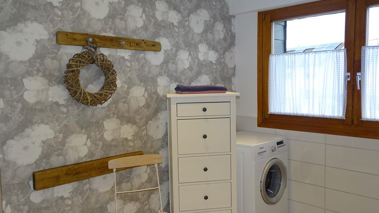 Ein modernes Badezimmer mit einem hellen, gemusterten Hintergrund und einem großen Fenster. Eine Waschmaschine steht neben einem weißen Schrank und einem Stuhl.