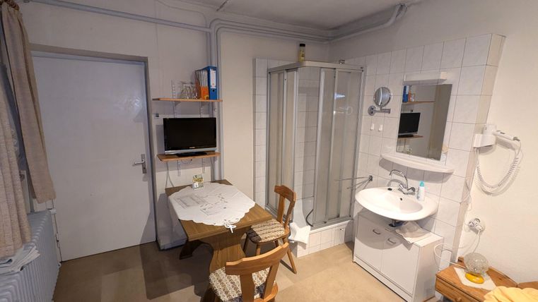Ein kleines Zimmer mit einem Tisch, zwei Stühlen und einem Waschbecken. In der Ecke befindet sich eine Dusche und ein Fernseher an der Wand.