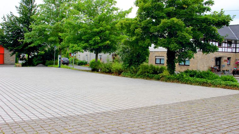 Ein leerer Platz mit einem Pflasterboden und einigen Bäumen. Im Hintergrund sind Gebäude zu sehen.