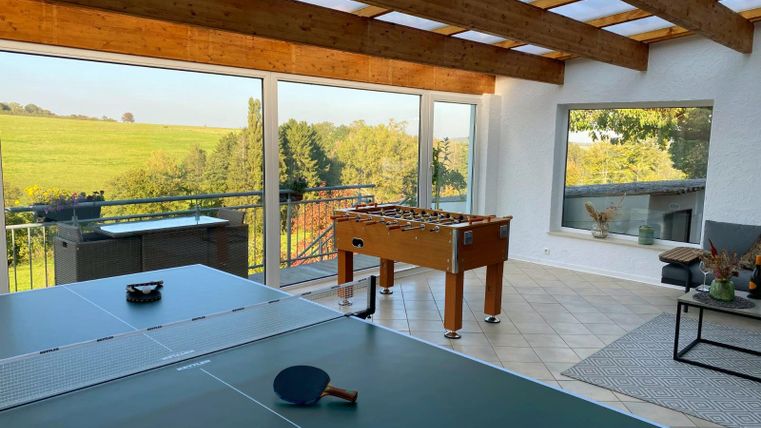 Ein heller Raum mit Tischtennisplatte und Tischfußball, umgeben von großen Fenstern. Im Hintergrund sind grüne Felder und Bäume sichtbar.
