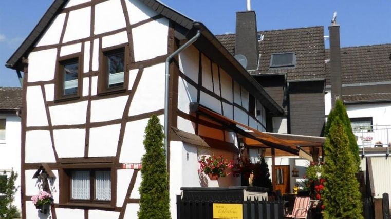 Ein charmantes Fachwerkhaus mit einem sonnigen Eingangsbereich. Vor dem Haus stehen grüne Pflanzen und bunte Blumen.