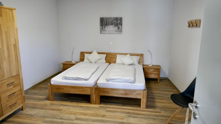 Ein einfaches, modernes Schlafzimmer mit zwei Einzelbetten und Holzmöbeln. Die Wände sind in einem neutralen Farbton gehalten, und ein Bild von einem Wald hängt über dem Bett.